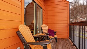 Una terraza de madera con dos sillas y una mesa, una puerta que da a un espacio interior, y una vista de la calle y árboles al fondo.