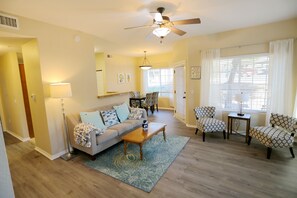 A living room with a sofa, coffee table, patterned chairs, a ceiling fan, and a window with blinds.
