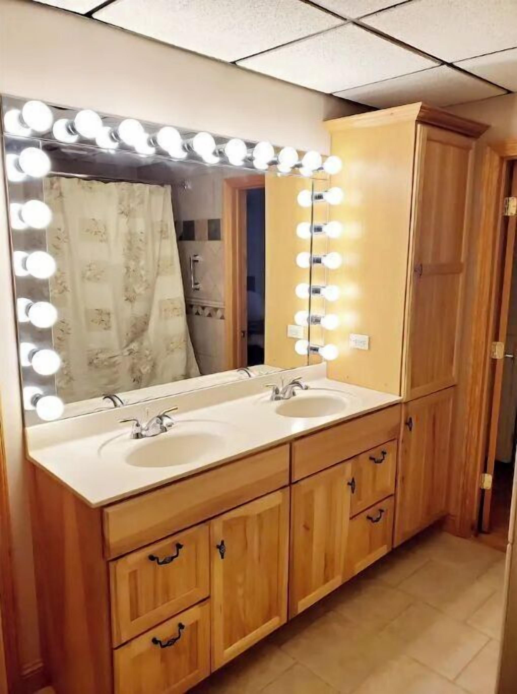 A bathroom with a vanity, two sinks, a mirror with lights, wooden cabinets, and a shower curtain.
