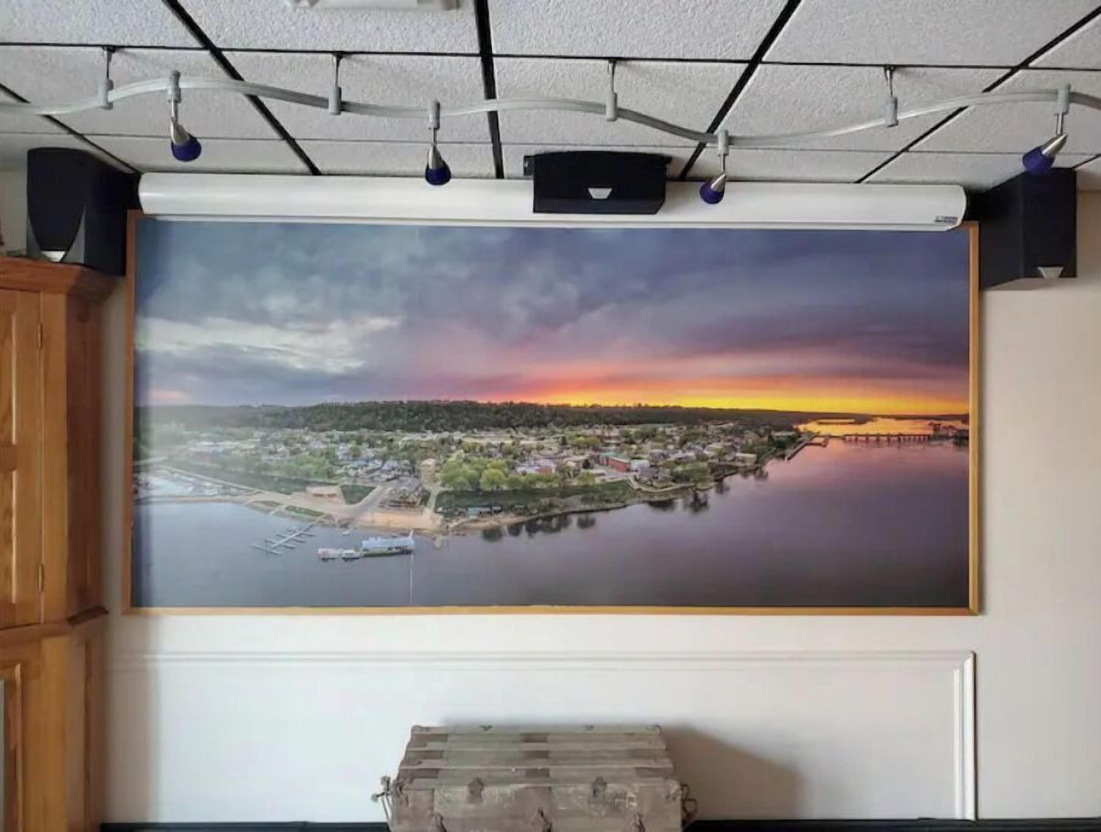 A large framed landscape painting of a sunset over a river with buildings on the opposite bank.