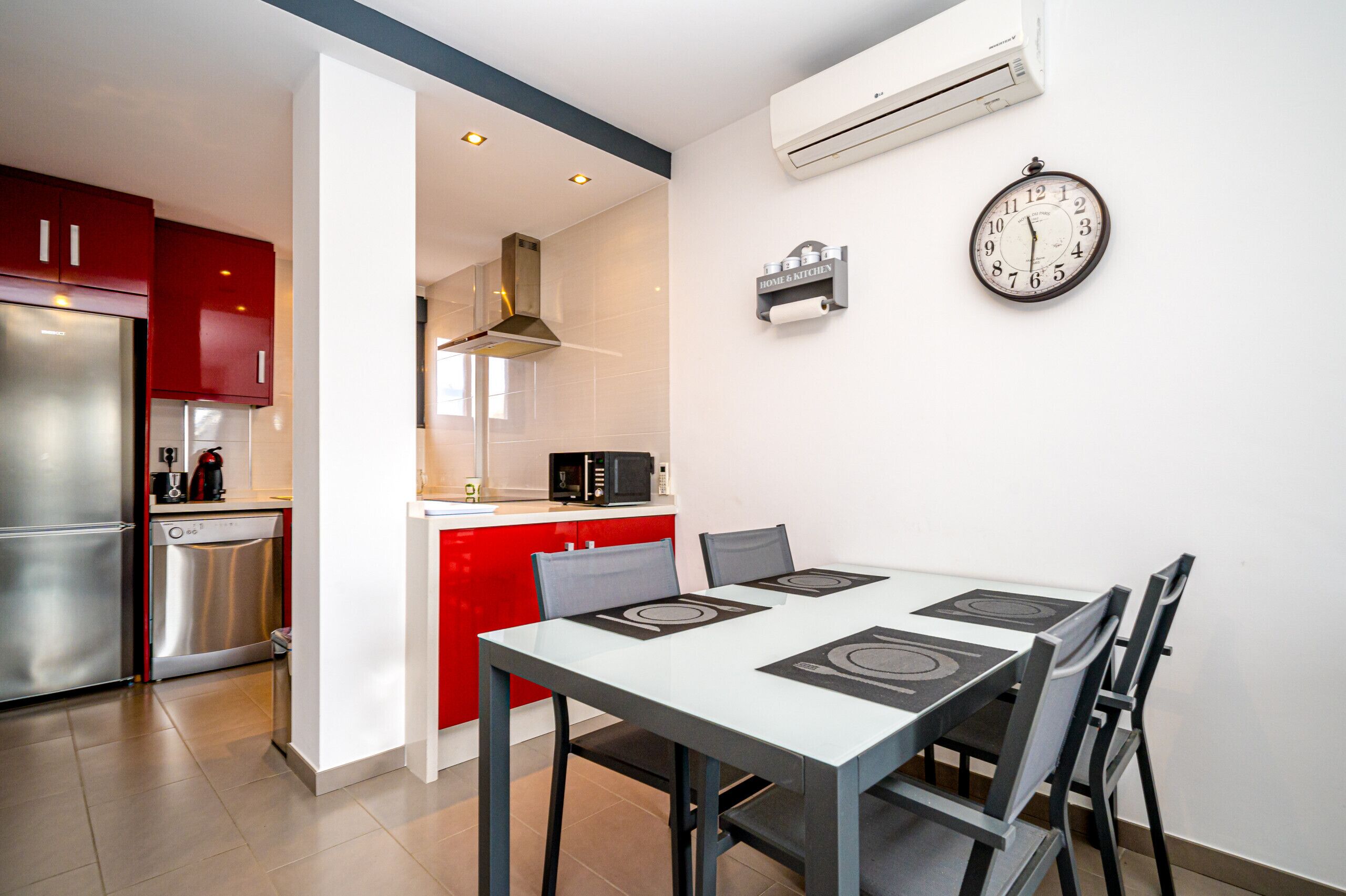 A modern kitchen with red cabinets, a stainless steel refrigerator, a dishwasher, and a microwave. There is a dining table with chairs and a clock on the wall.