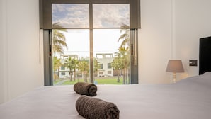 Habitación de hotel con una cama grande, dos toallas enrolladas, una lámpara y vista a un edificio y palmeras a través de una ventana.