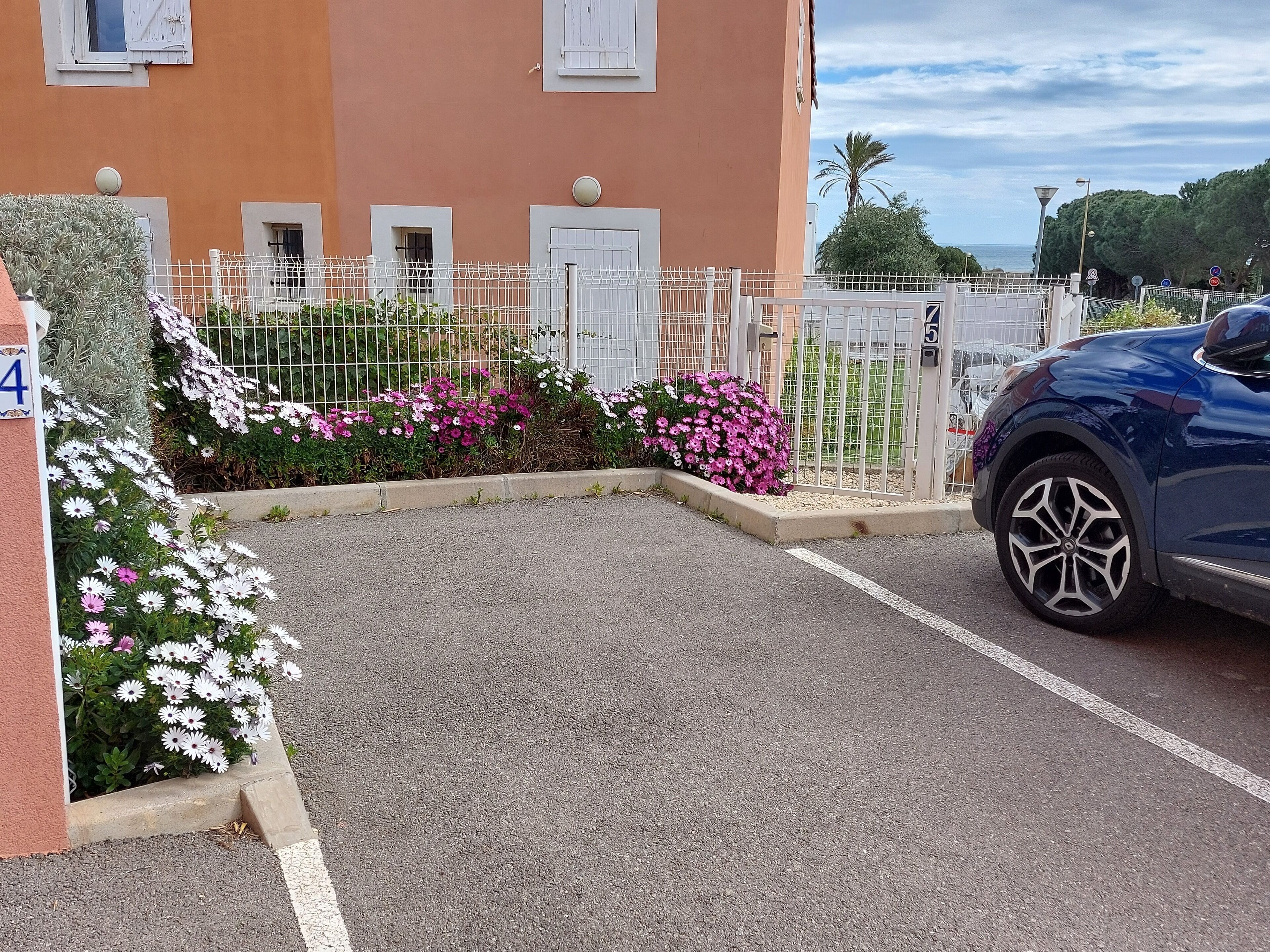 Un área de estacionamiento con un coche azul, un edificio con el número 4 y una cerca blanca adornada con flores.