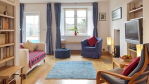 A cozy living room with a sofa, armchair, ottoman, and a TV. There is a bookshelf, a window with curtains, and a small table with fruit.