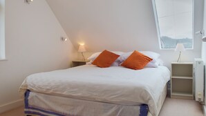 A bedroom with a sloped ceiling, a bed with white bedding and orange pillows, a bedside table with a lamp, and a window with a view of the sky.