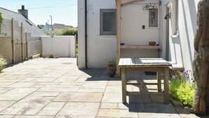 A small, white building with a wooden table and bench in the foreground, a small window, and a stone-paved courtyard.