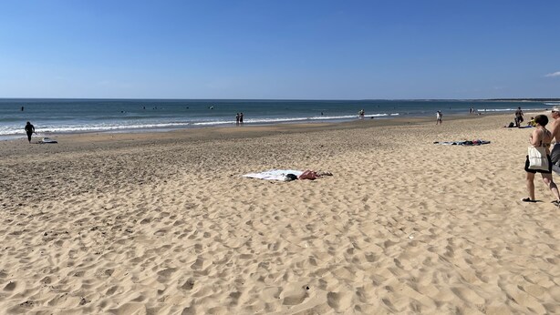 A beach with people walking and standing, and a few umbrellas on the sand.