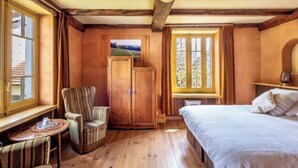 Une pièce avec un plancher en bois, un lit, une chaise, une petite table, une armoire et une fenêtre donnant sur des arbres.