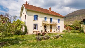 Une maison à deux étages avec un toit recouvert de tuiles rouges, un balcon et un jardin agrémenté d’une table et de chaises.