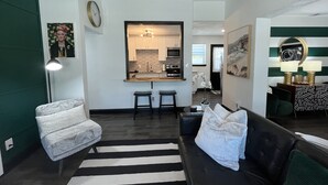 A modern living room with a black leather sofa, a patterned armchair, and a striped rug. A kitchen area is visible in the background with white cabinets and a large mirror above the counter.