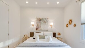 Une chambre à coucher bien aménagée, avec un grand lit, deux tables de chevet, une œuvre d’art accrochée au mur et une fenêtre avec des stores.
