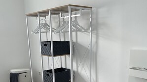 A modern room with a white wardrobe featuring multiple shelves and hanging space, a small trash bin, a bedside table with a lamp, and a grey sofa.