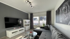A modern living room with a flat-screen TV, a white entertainment unit, a grey sofa, a glass coffee table, and a large window with curtains.