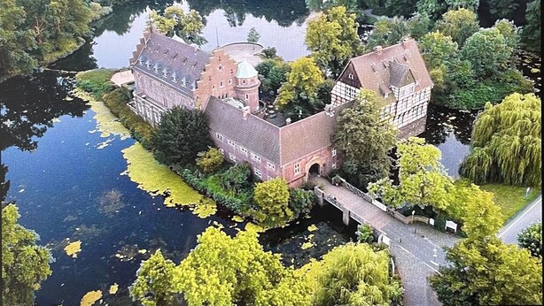 A historic building with a moat, surrounded by lush greenery and a calm body of water.
