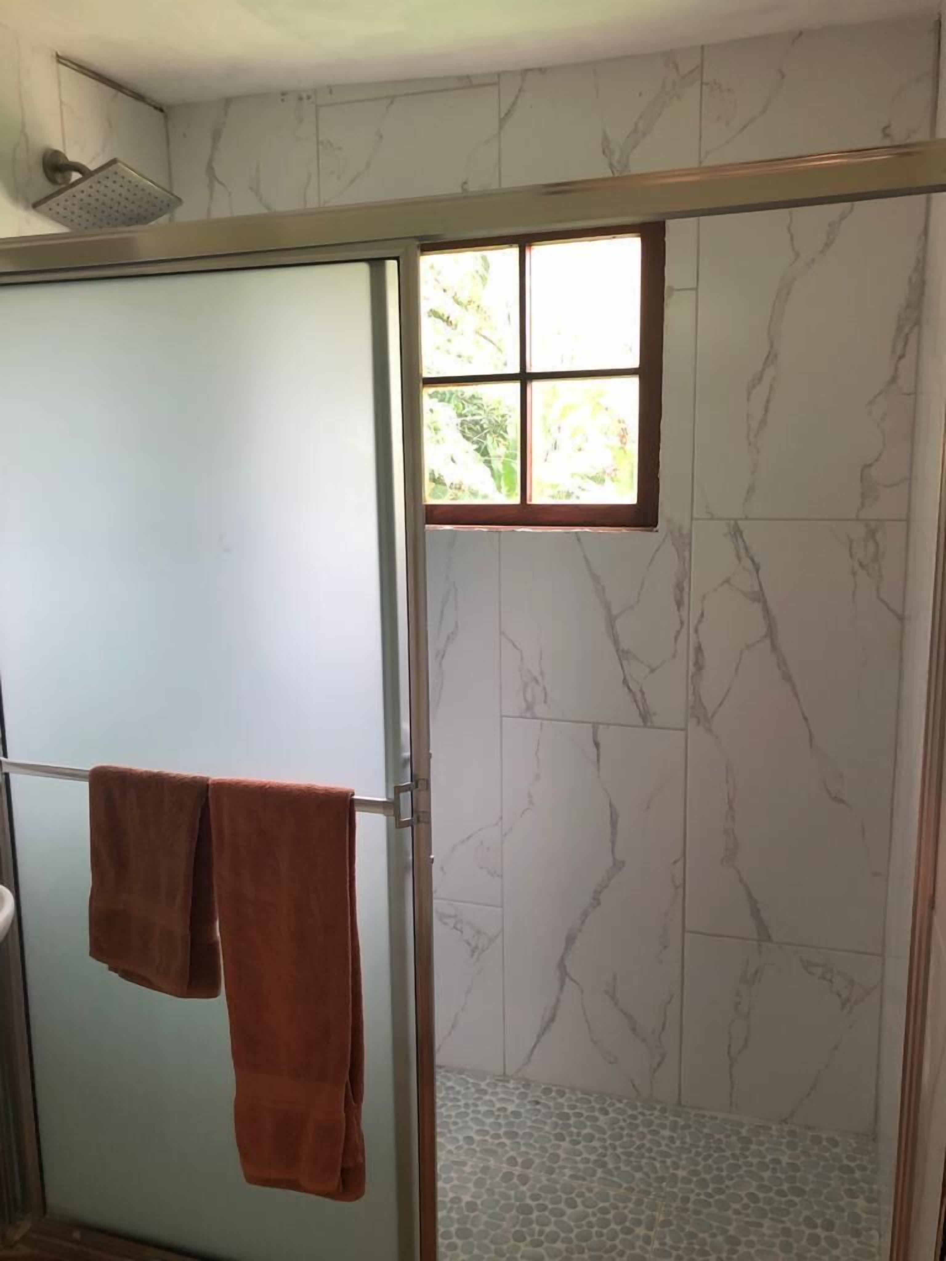Baño con zona de ducha, paredes de mármol y ventana con vistas a la vegetación.