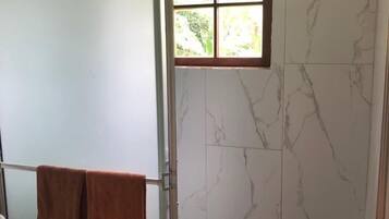 Baño con zona de ducha, paredes de mármol y ventana con vistas a la vegetación.