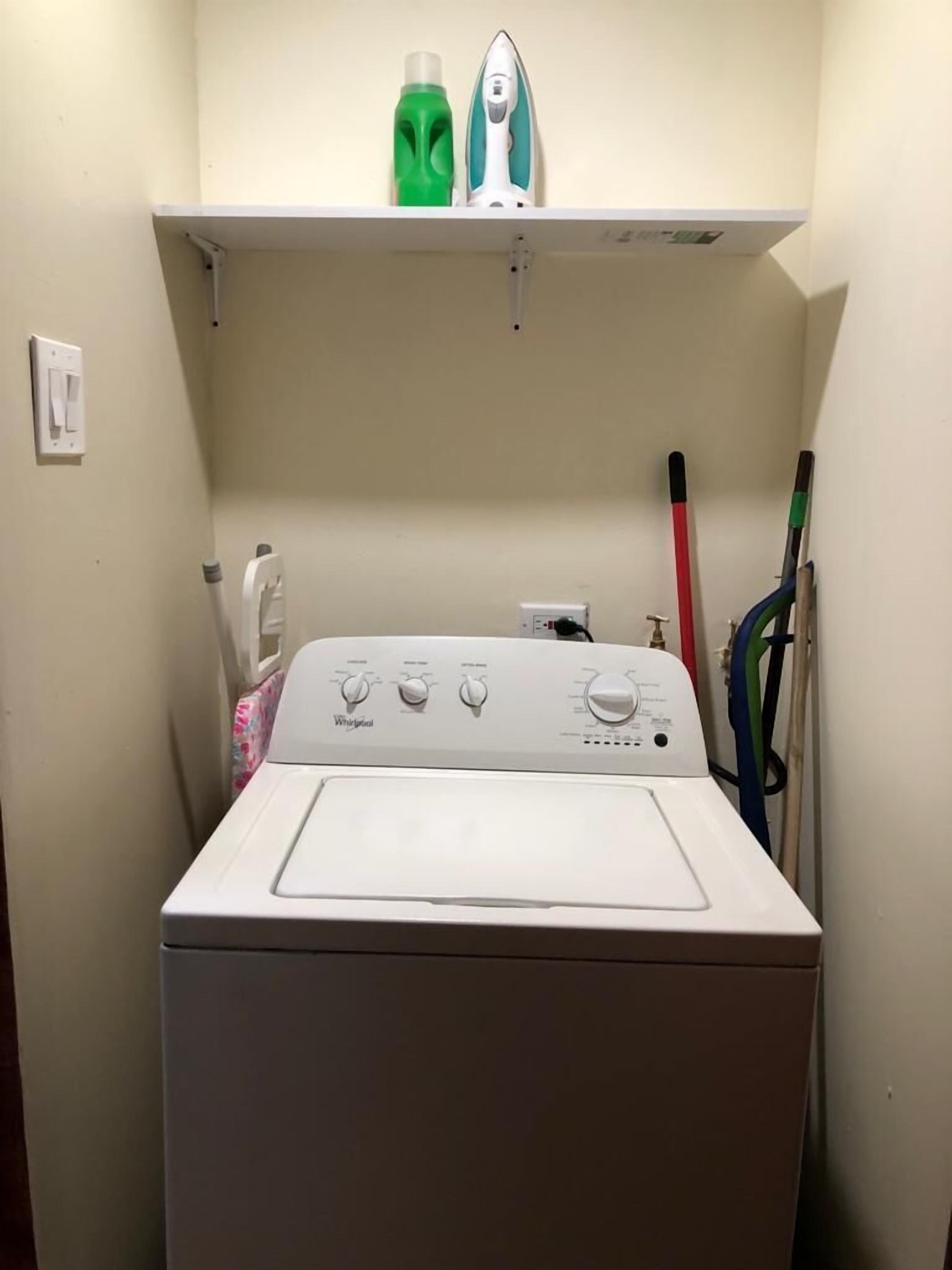 Una lavadora Whirlpool de carga frontal con un panel de control, una botella verde y un planchador en un estante superior.