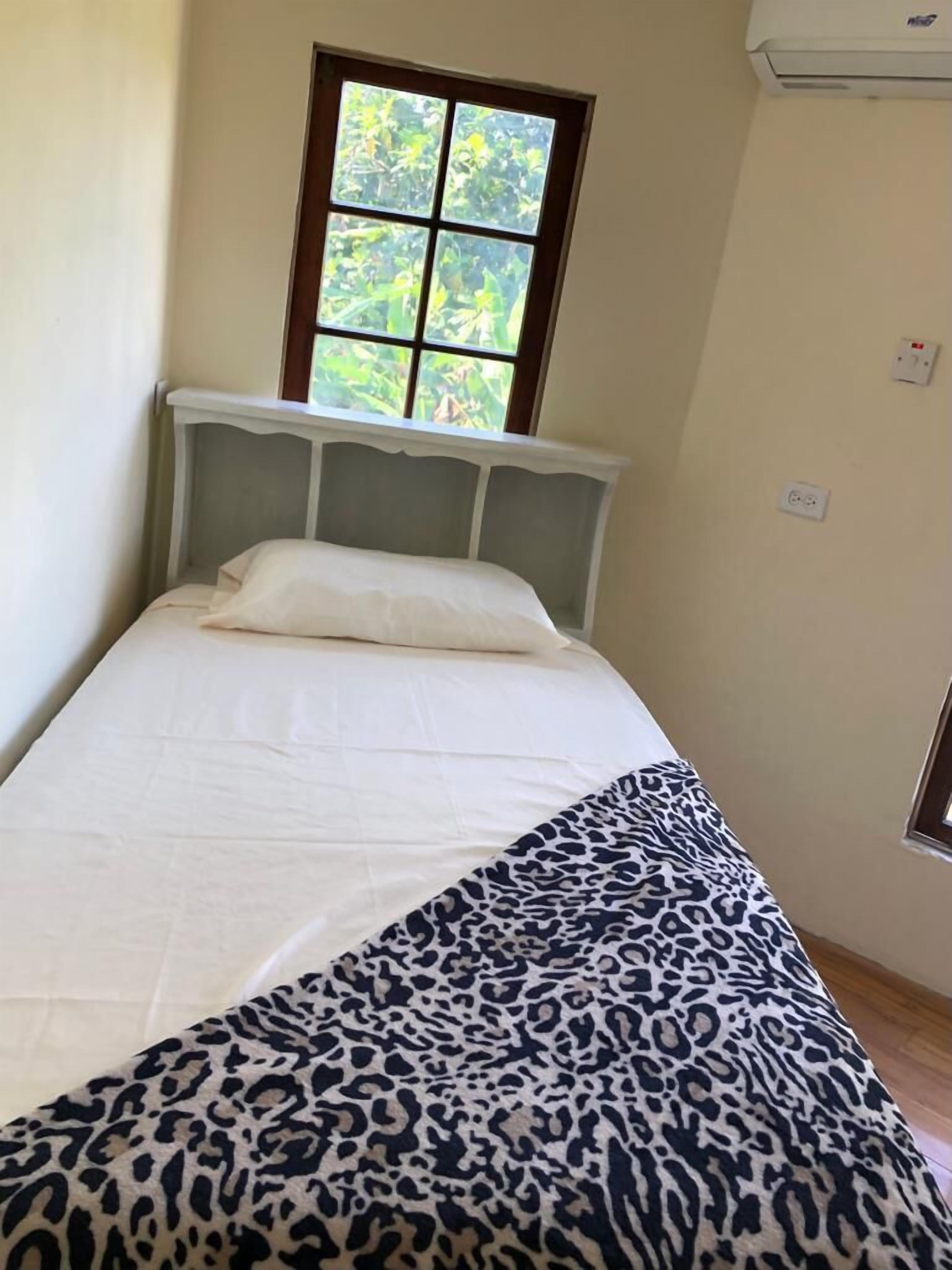 Un lit simple avec un matelas blanc et une couverture à motifs léopard. Il y a une fenêtre au-dessus du lit, donnant sur un paysage verdoyant.