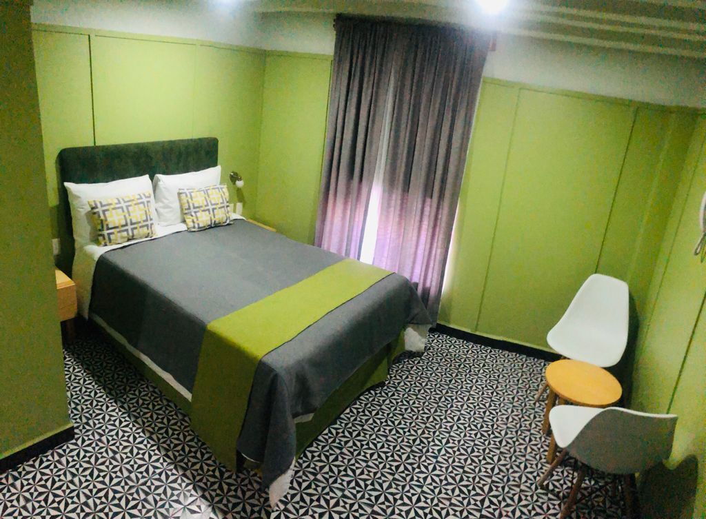 Hotellihuone, jossa on sänky, yöpöytä, tuoli, pöytä ja verho.