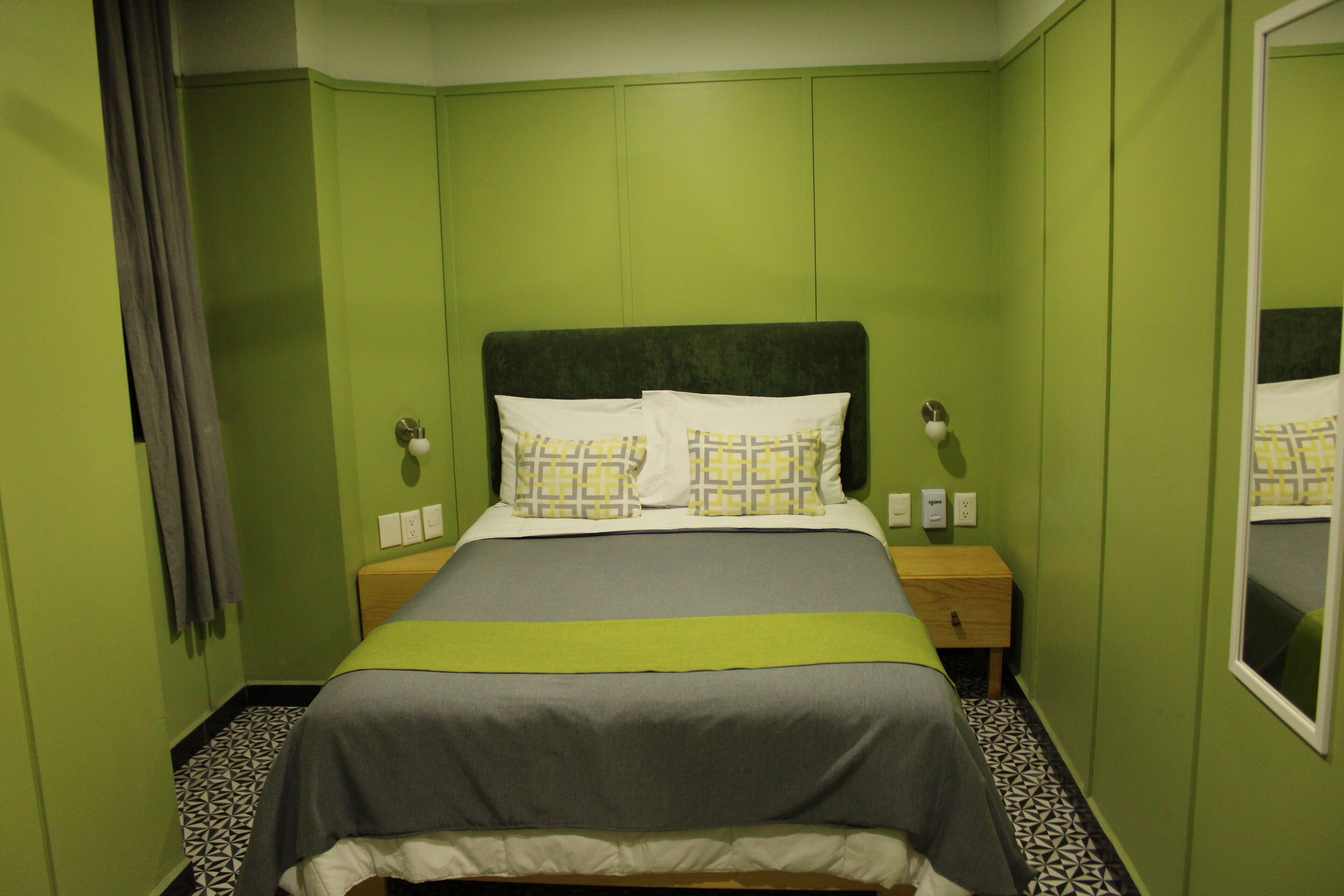 A neatly made bed with a grey and white bedspread, two pillows, and a green accent. There is a wooden nightstand with a lamp and a power outlet. The room has green walls and a patterned floor.