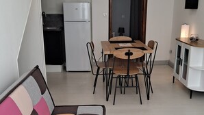 Een moderne woonkamer met een kleurrijke bank, een eethoek met een tafel en stoelen, een keuken met een koelkast, en een klein kastje.