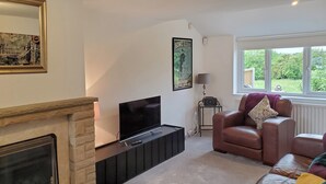 Ein Wohnzimmer mit Kamin, Fernseher, Ledersofa und einem Fenster mit Blick auf Grünflächen.