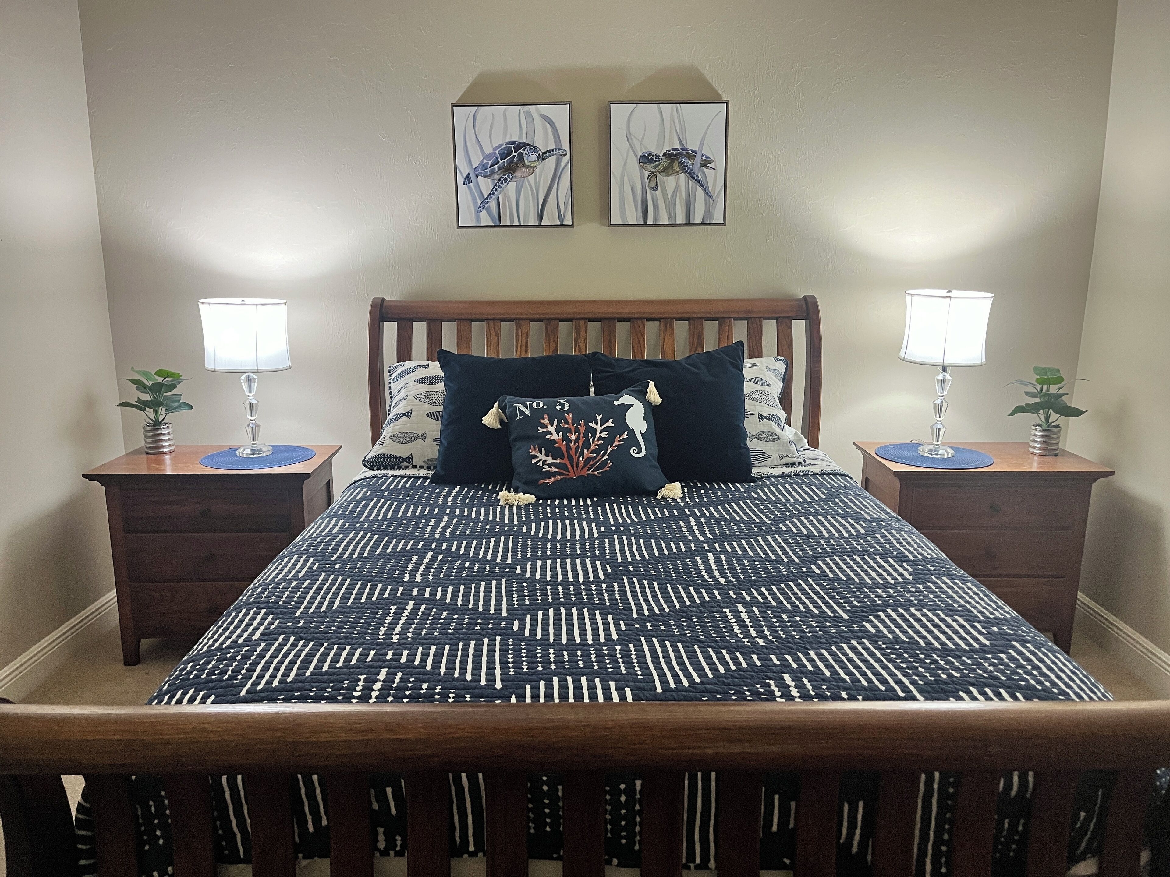A neatly made bed with a patterned bedspread, two wall-mounted lamps, and two framed artworks above the bed.