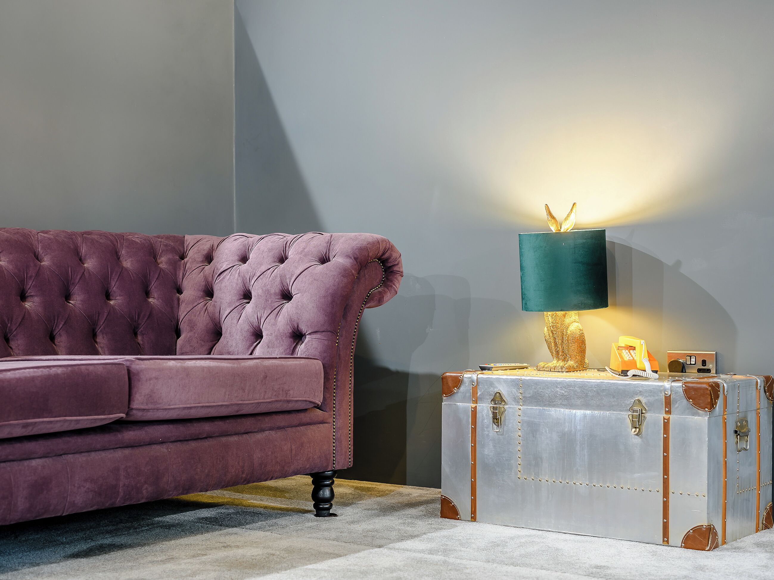 Een stoffen paarse bank, een lamp in vintage stijl, en een metalen koffer met slot.