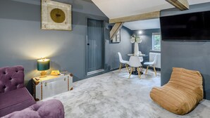 Un salon moderne avec un plafond haut, des poutres apparentes, une table ronde avec des chaises, un téléviseur et un coin salon confortable.