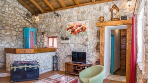 Ein gemütliches Wohnzimmer mit Steinwänden, Holzbalkendecke, Fernseher, einem grünen Sessel und einem kleinen Tisch mit Weingläsern.