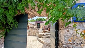 Ein Steinweg führt zu einem Steinhaus mit Tisch und Stühlen unter einem violetten Sonnenschirm.