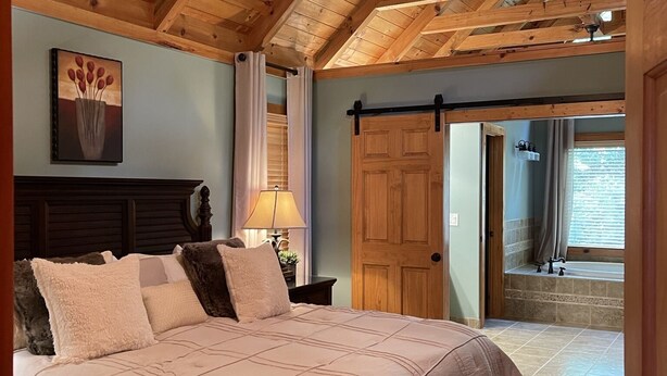 Un dormitorio con techo de madera, una cama con edredón color crema, una mesita de noche con lámpara y un cuadro en la pared.
