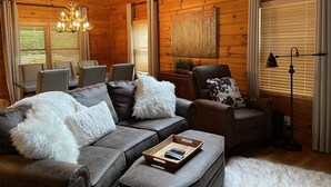 Un acogedor interior de cabaña con techo de madera, un sofá gris con almohadones blancos mullidos, un sillón de cuero marrón y un comedor con mesa y sillas.