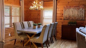 Interior de una cabaña de madera con una mesa de comedor, sillas, una lámpara de araña y un cuadro en la pared.