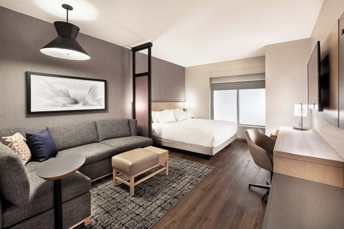 Een moderne hotelkamer met een grijze bank, een bed met witte lakens, een bureau met stoel, een groot raam en een wandgemonteerde televisie.