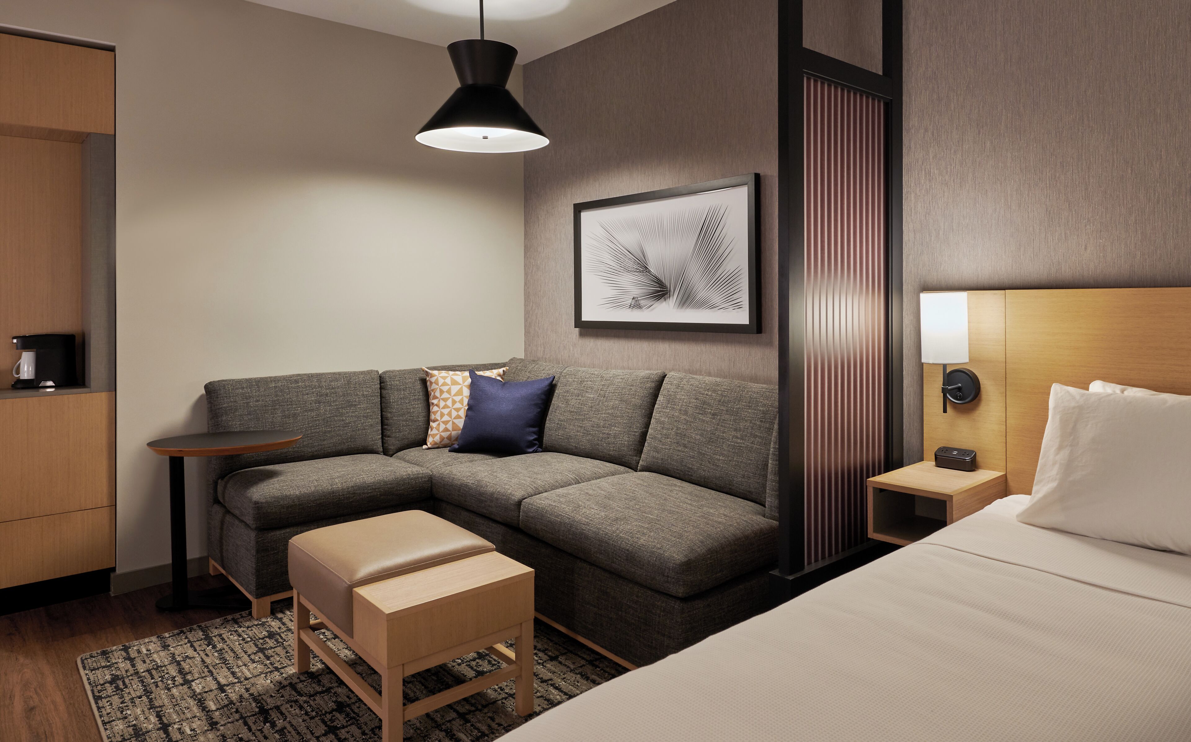 Una habitación de hotel moderna con un sofá seccional gris, un banco de madera, una mesita de noche pequeña, una lámpara de pared y una obra de arte enmarcada.