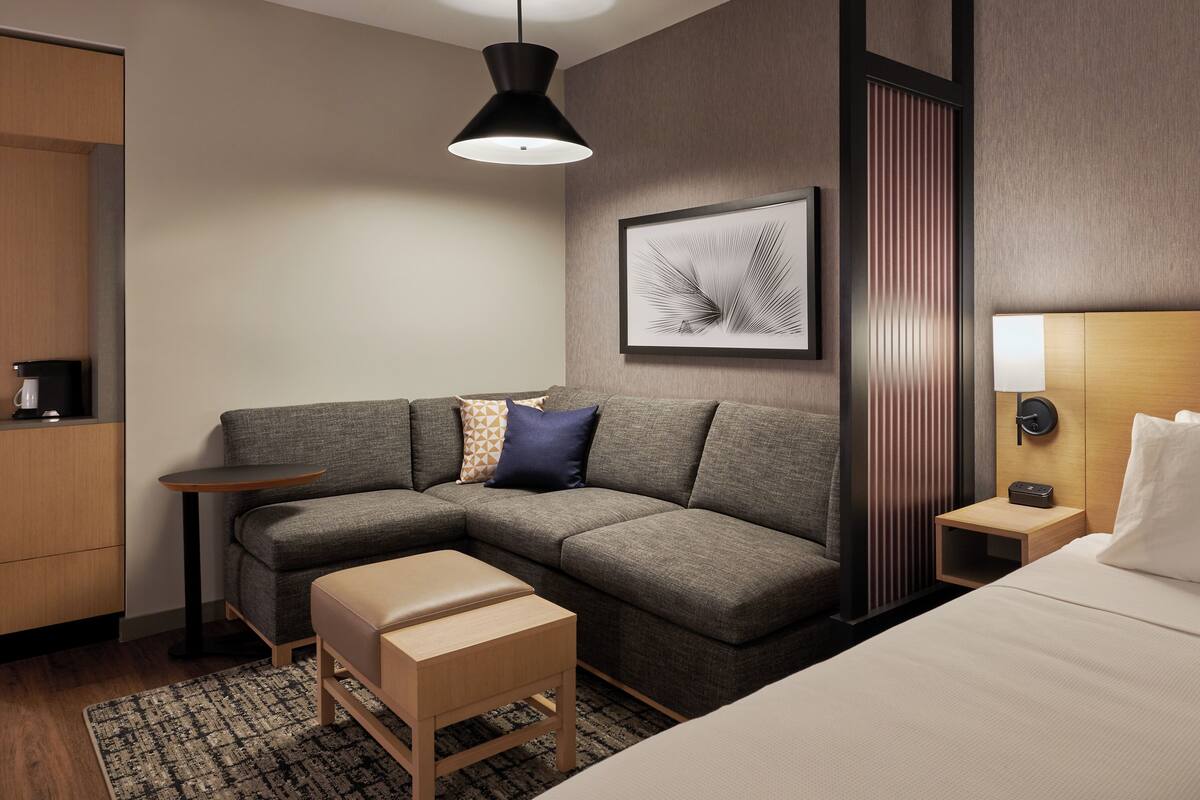 Een moderne hotelkamer met een grijze modulaire bank, een houten bankje, een klein nachtkastje, een wandlamp en een lijstwerk met kunst.
