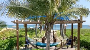 En træpergola med en hængekøje og bænk, omgivet af frodig vegetation og kig til havet.