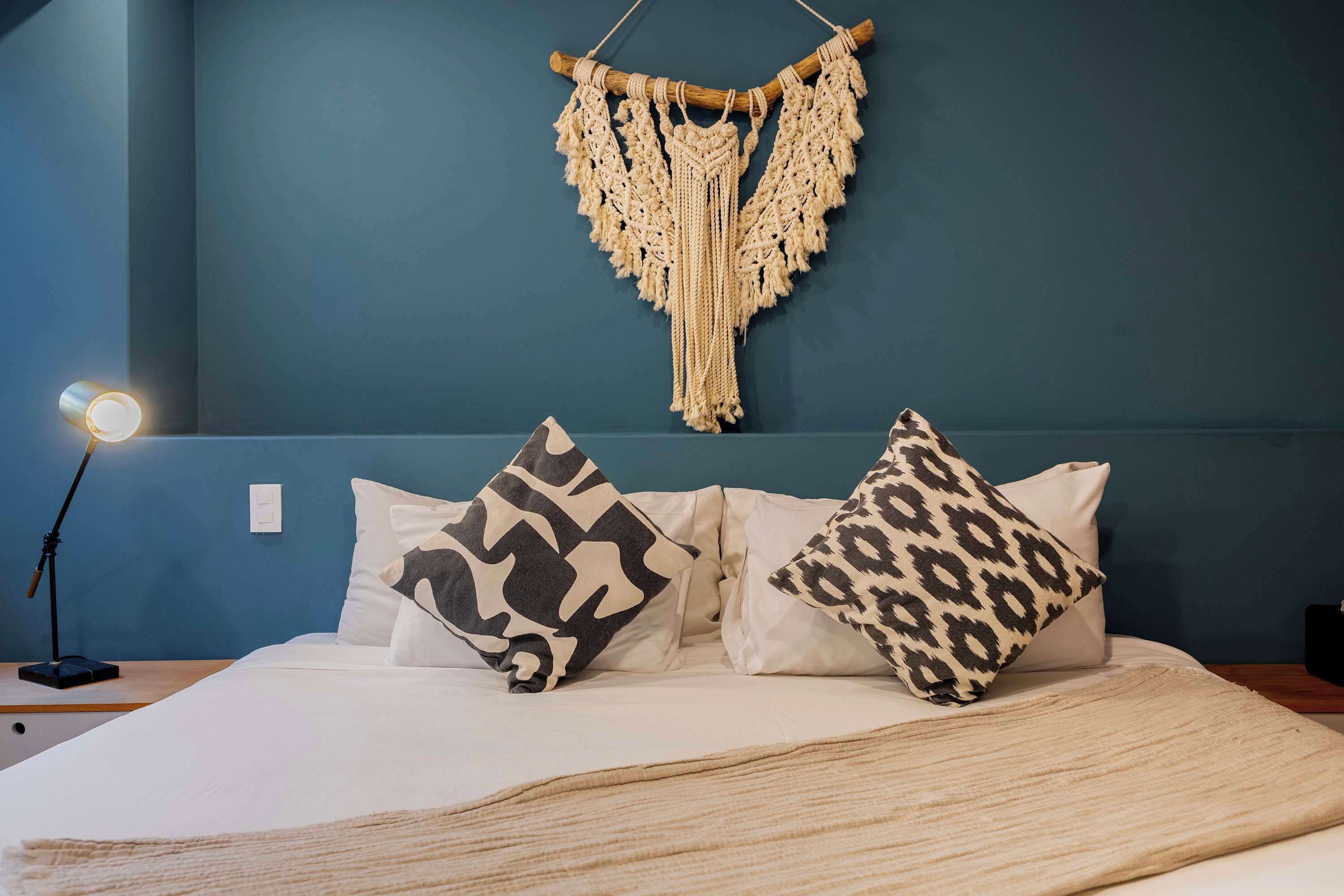 A bed with white bedding, two patterned pillows, a macrame wall hanging, and a bedside lamp.