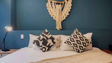 A bed with white bedding, two patterned pillows, a macrame wall hanging, and a bedside lamp.