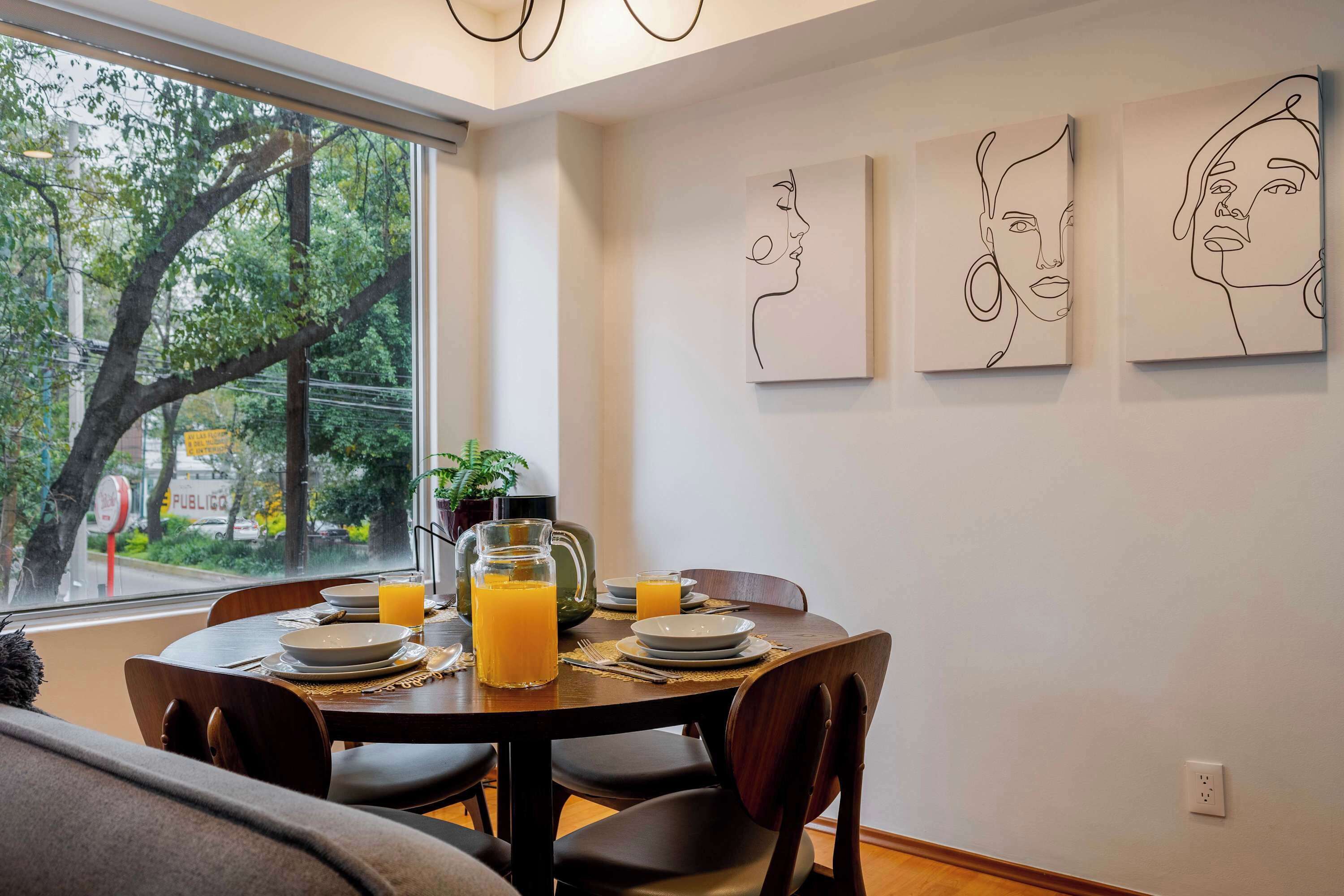 Uma área de refeições com uma mesa posta para dois, uma jarra de suco de laranja e vista para árvores.