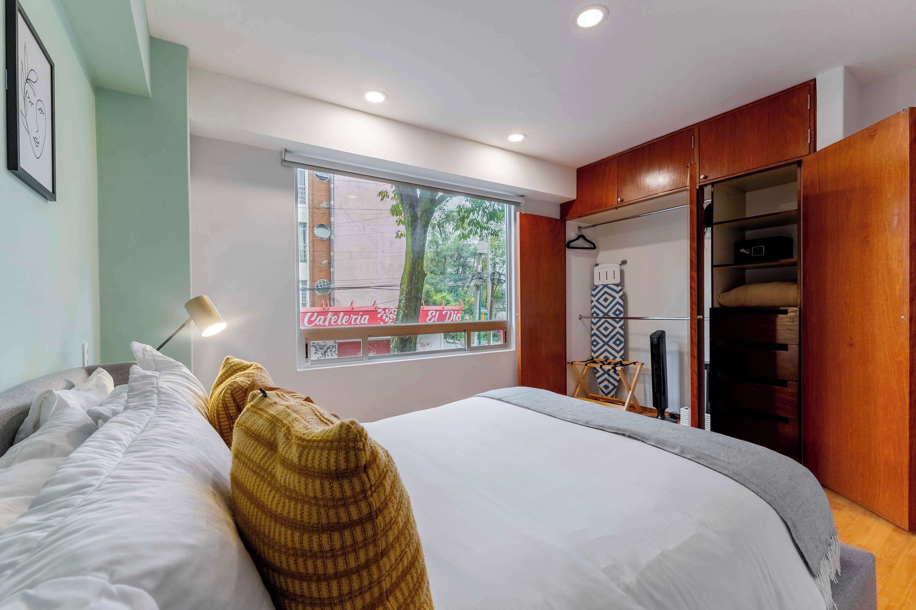 Quarto com uma cama, janela com vista para um edifício, guarda-roupa e mesa de cabeceira com abajur.