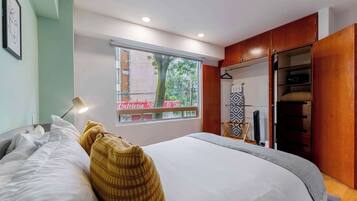 Quarto com uma cama, janela com vista para um edifício, guarda-roupa e mesa de cabeceira com abajur.