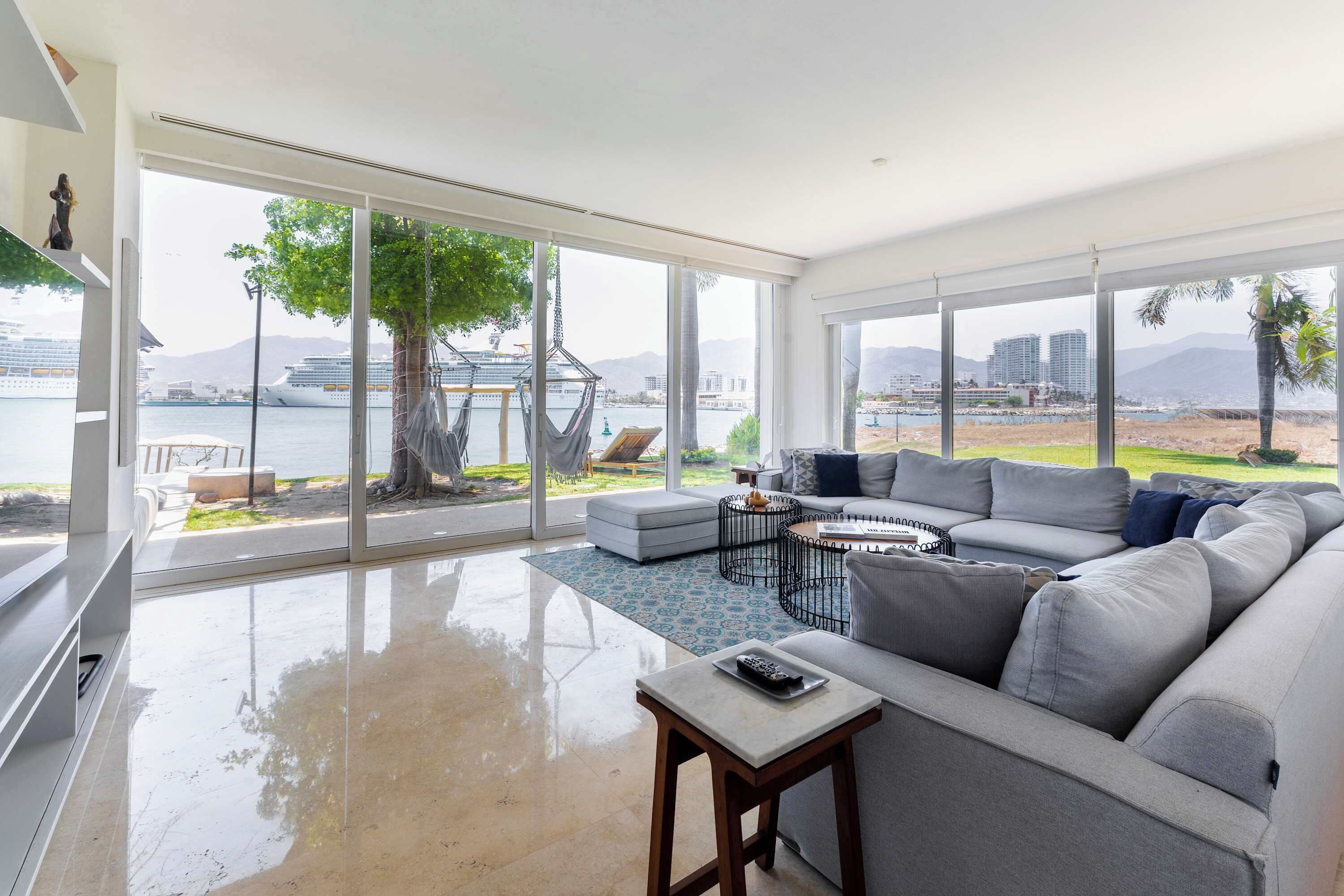 A spacious living room with a large window, a grey sofa, a small table, and a hammock.