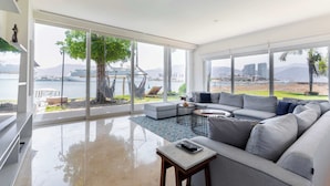 A spacious living room with a large window, a grey sofa, a small table, and a hammock. - Casa Luna del Mar - Private Pool & Beach, FastWifi (Puerto Vallarta)