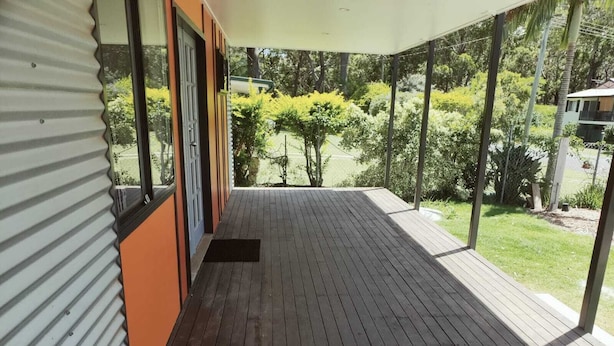 Una terraza de madera con barandilla metálica, una puerta que da a un espacio cubierto y vistas a un jardín con diversas plantas.