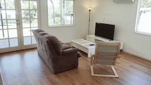 Una sala de estar amplia con un sofá marrón, un televisor sobre un mueble bajo, una silla de madera y ventanas grandes.