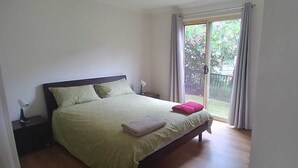 Un dormitorio con cama, mesita de noche, ventana con cortinas y vista al verdor exterior.