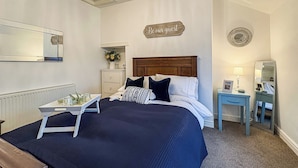 A neatly arranged bedroom with a bed, bedside table, dresser, mirror, and a sign on the wall.
