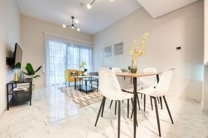 A modern living room with a dining area, a flat-screen TV mounted on the wall, and a large window with sheer curtains. - Stylish 1 BR + Pool & World Class Gym (Dubai)